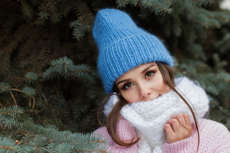 A woman with a winter hat, scarf and sweater on standing in front of a pine tree