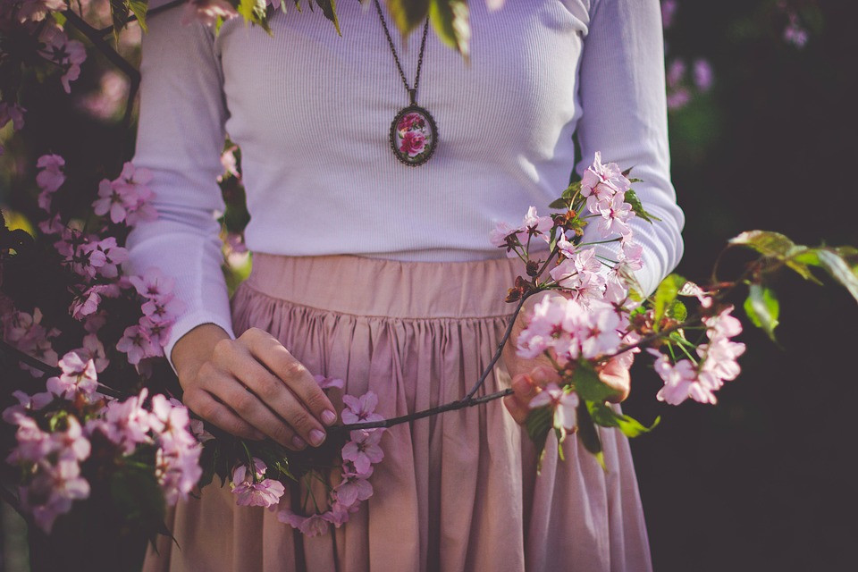 A woman wearing a big necklace standing next to a pink flower tree holding a branch
