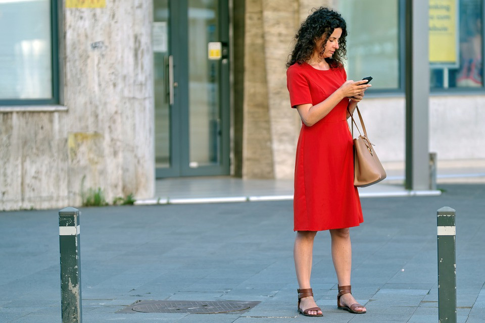 A woman standing on the sidewalk in a red dress, looking at her phone with a beige purse
