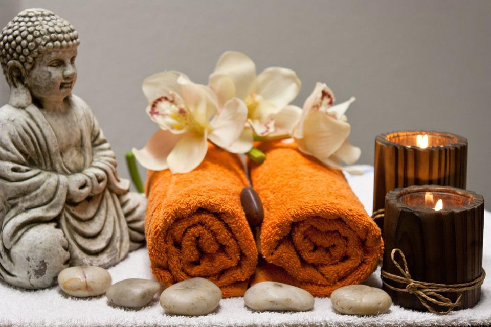 A Buddha statue, orange rolled towels, flowers, stones and candles on a towel