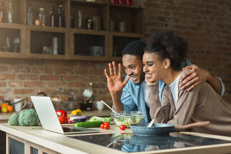 A couple enthusiastically participates in an online cooking class, waving at their laptop screen.