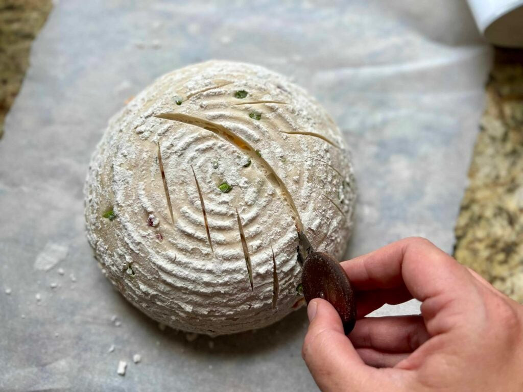 A hand scoring a sourdough loaf with a bread lame, creating a decorative pattern on the dough's surface before baking.