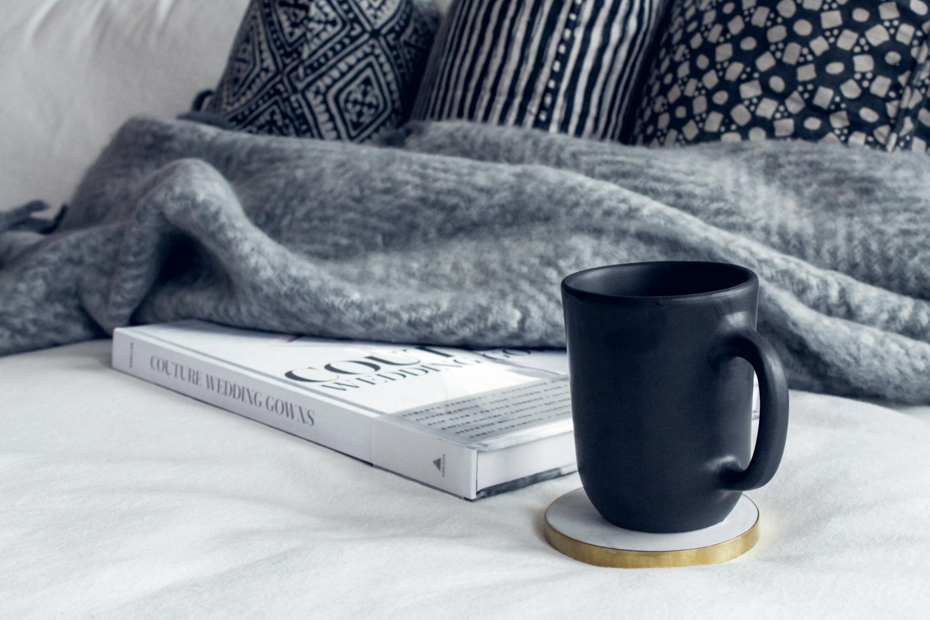 black ceramic mug on round white and beige coaster on white textile beside book and throw blanket with pillows