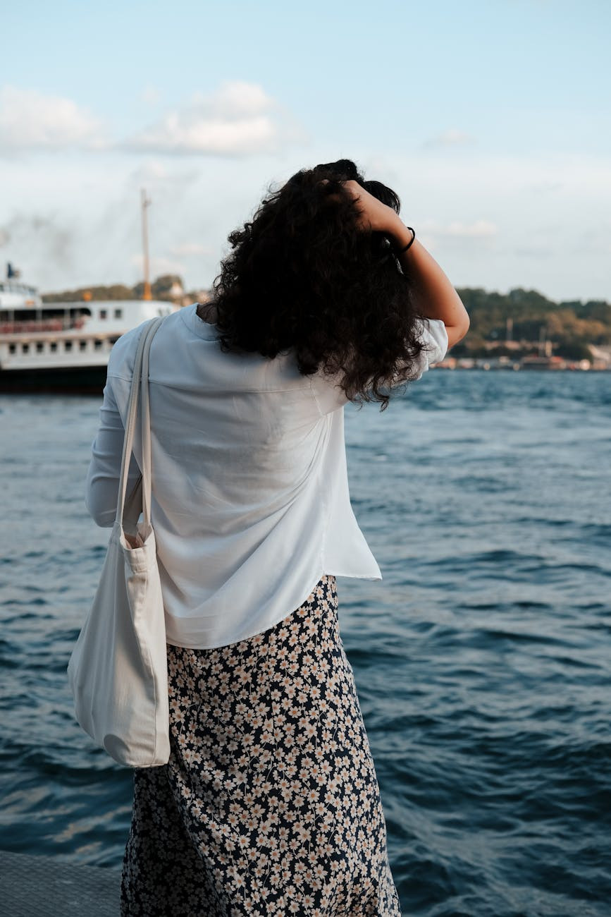 brunette woman fixing hair standing at sea with a canvas tote bag on her shoulder