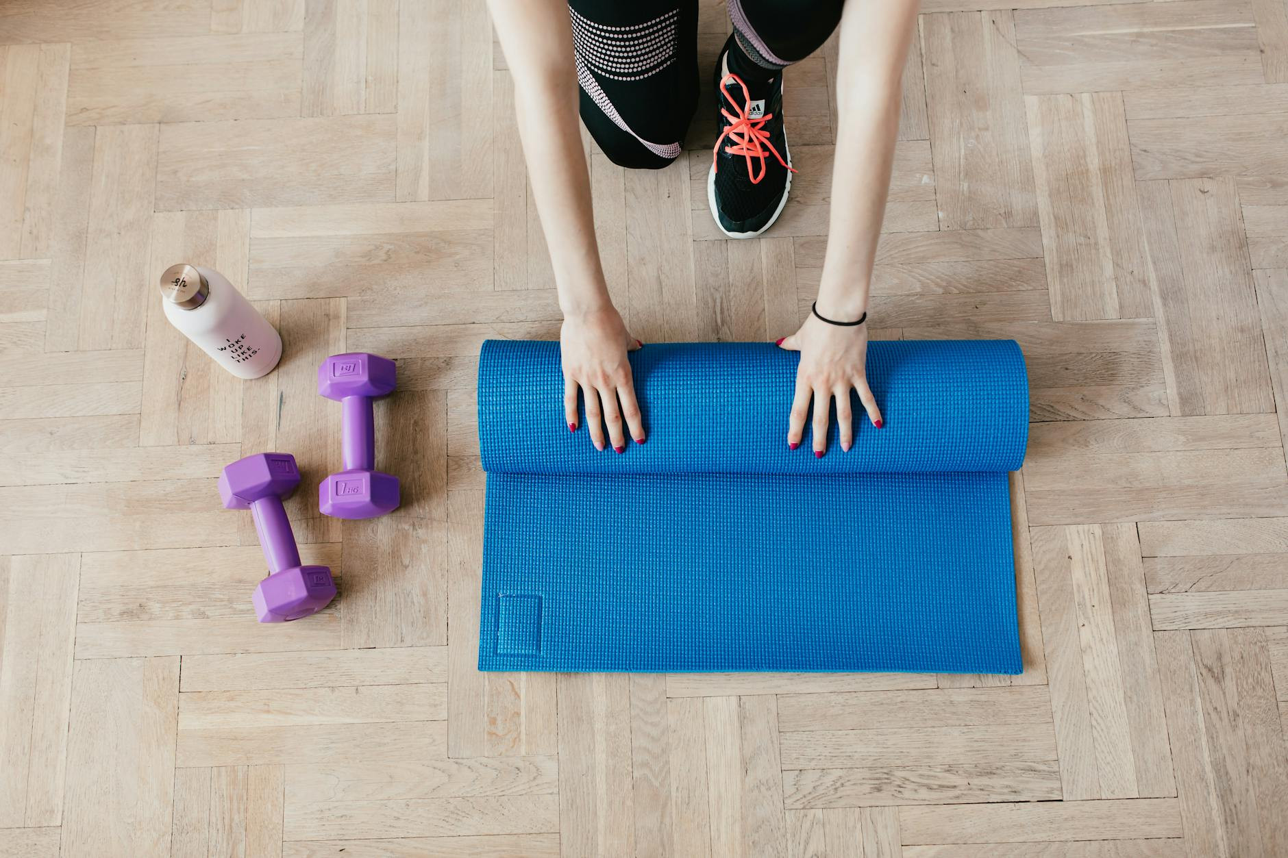 crop sportswoman unfolding sport mat on wooden floor with hand weights and custom water bottle next to it